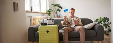 Yakışıklı erkek manken resmi, bavullu turist, uçakta iki biletini gösteriyor, cep telefonu uygulaması kullanıyor, kanepeye oturuyor. Turizm ve seyahat kavramı