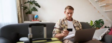 Neşeli bir adamın portresi, tatile çıkıyor, yurtdışına seyahat planları yapıyor, dizüstü bilgisayarı açık, oturma odasında bavuluyla oturuyor, bir otel rezervasyonu yaptırıyor..