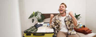 Heyecanlı kulaklıklı genç bir adam, tatile çıkıyor, eşyaları bavula koyuyor, kulaklıkla müzik dinliyor, bagajının yanına oturuyor. Turizm ve yaşam tarzı kavramı