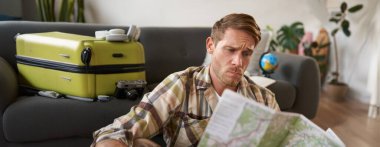 Endişeli, kafası karışmış, haritaya bakan, rotayı incelerken kaşlarını çatan, bavulla oturan, tatile çıkan bir adamın portresi..