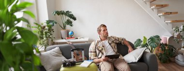 Yakışıklı genç adamın portresi, bavul ve iki bilet yanında dijital tabletle oturan turist, tatili için internetten bir şeyler ayırtıyor, tatil hazırlıyor..