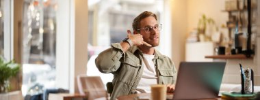 Gözlüklü genç yakışıklı bir adamın portresi, kafede çalışan iş adamı, dizüstü bilgisayarla oturan, telefonla el hareketi gösteren, servisini tanıtan.