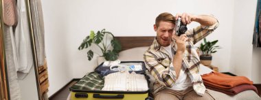 Yakışıklı mutlu adamın portresi, analog kamerasını tutuyor, açık bavulun yanında oturuyor, tatil için eşyalarını topluyor, yurt dışına seyahat ediyor, tatile hazırlanıyor..