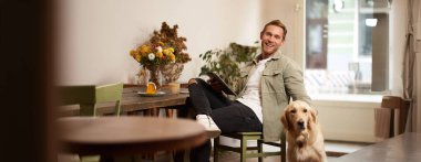 Yakışıklı genç bir adamın portresi, kafe ziyaretçisi, köpeğiyle oturuyor Golden Retriever 'ı okşuyor ve dijital tablet, yaşam tarzı ve evcil hayvan dostu kamusal alanlar hakkında haberler okuyor..