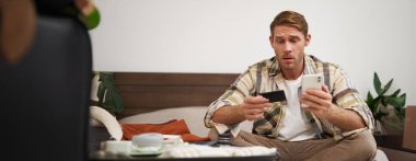 Genç bir adamın portresi, internetten ödeme yapan bir turist, kredi kartı ve cep telefonu uygulaması kullanıyor, bavulla yatakta oturuyor, internetten sipariş veriyor, tatil biletleri alıyor..