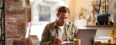 Gözlüklü, kafede oturan, notlar alan, çevrimiçi kurslara katılan, uzaktan kumandalı bir kafeden öğrenen odaklanmış bir genç resmi..