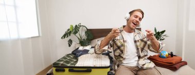 Sarışın, kulaklıklı yakışıklı bir adam, bavul ve kıyafetlerle yatakta oturuyor, tatil için hazırlanırken kablosuz kulaklıkla müzik dinliyor, seyahate çıkıyor..