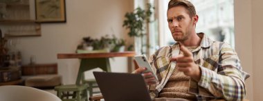 Ciddi yüzlü bir adamın portresi, laptoplu ve akıllı telefonlu bir kafede oturuyor, seni uyarıyla işaret ediyor, azarlıyor, onaylamayarak kaşlarını çatıyor..