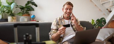 Kredi kartlı genç bir adamın portresi, oturuyor ve dizüstü bilgisayar kullanıyor, bir telefon görüşmesi yapıyor, otel ya da bilet ayırtıyor, telefonla satın alımını onaylıyor, yanında bavulu var..