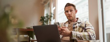 Laptop ve cep telefonuyla oturan kafeterya ziyaretçisinin portresi kafası karışmış ve kameraya şaşırmış görünüyor. Yaşam tarzı ve kamusal alan kavramı