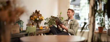 Yakışıklı, gülümseyen genç bir adamın portresi, kahve dükkanında oturuyor, dijital tablet kullanıyor, kafeden biriyle görüntülü sohbet ediyor, kablosuz kulaklık takıyor..