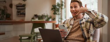 Komik, yakışıklı bir kafe ziyaretçisinin portresi, dizüstü bilgisayarı ve akıllı telefonu olan genç adam, cep telefonu işaretini gösteren ve kameraya gülümseyen, onu aramak isteyen, telefonla soran..