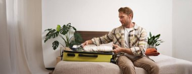 Yolculuk için bavulunu toplayan genç adamın portresi, tatile gitmek, yatağa oturmak ve giysilerini bavula koymak, uçuşu için hazırlanmak..