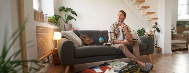 Tatile giderken yanında ne götüreceğini düşünen bir adamın portresi, elinde defter olan, düşünceli yüzlü eşyaların listesi, tatile çıkmaya hazırlanan, kıyafetlerle valizin yanında oturan..