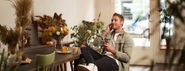 Gündelik giysiler içinde genç yakışıklı bir adamın portresi, kafede oturuyor ve en sevdiği şarkıyı seviyor, kablosuz kulaklıkla müzik dinliyor, kulaklıkla rahatlatıcı melodileri dinliyor, akıllı telefon tutuyor..