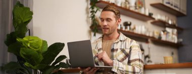 Bilgisayarlı yakışıklı bir gencin portresi, kafede oturuyor, ekrana bakıyor, iş yapıyor, serbest çalışıyor, uzaktan çalışma alanı var..