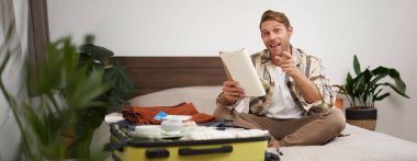 Mutlu, karizmatik genç bir adamın portresi, tatil için heyecanlı, elinde not defteri, tatilde giyecek dolu bavul, kamerayı işaret etmek ve gülümsemek..