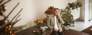 Ofisteki yakışıklı mutlu adamın portresi, ortak çalışma alanında oturan, kameraya bakan, gülümseyen ve gülen, cep telefonuyla konuşan, akıllı telefon kullanan, müşterinin telefonuna cevap veren adam..