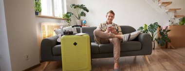 Genç turistin portresi, koltukta bavullu bir adam, tatile hazırlanıyor, akıllı telefonla oturuyor, cep telefonu uygulamasından otel ve uçak biletleri alıyor. Turizm ve seyahat kavramı