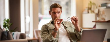 Adamın portresi şaşkın görünüyor, gözlüklerine bakıyor, mavi gözlüklere bakıyor, laptopla kafede oturuyor, uzaktan bilgisayarla çalışıyor..