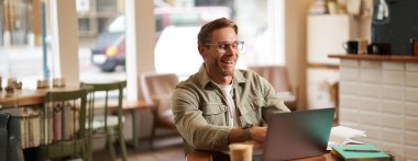 Genç dijital göçebe imajı, gözlüklü adam kafede oturuyor, kahvecide çalışıyor, iş yerinde dizüstü bilgisayar kullanıyor, gözlük takıyor, içkisini içiyor..