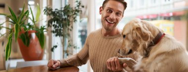 Gülümseyen genç adam köpeğinin patisini elinde tutuyor ve mutlu görünüyor. Hayvan dostu kafe ve serbest çalışma alanı kavramı.