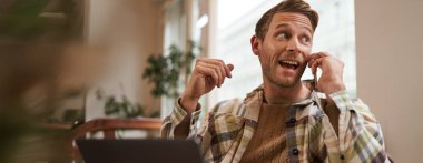 Serbest çalışma, dijital göçebe ve çalışma alanı kavramı. Cep telefonlu yakışıklı bir gencin portresi, laptopuyla kafede oturuyor, proje üzerinde çalışıyor, uzaktan çalışıyor, pencereden dışarı bakıyor..