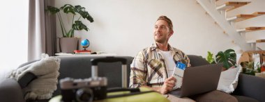 İnternetten uçuşunu kontrol eden, dizüstü bilgisayara bakan, elinde biletlerle evde bavulla oturan neşeli genç adamın portresi..