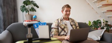 Mutlu genç adamın portresi iki kişilik tatil rezervasyonu, uçak biletleri, dizüstü bilgisayara bakması, oturma odasında bilgisayarla oturması. Seyahat ve turizm konsepti