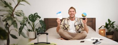 Neşeli, mutlu bir turistin portresi, uçak biletleri ve küreyle oturan bir adam, dünyayı dolaşmak için bavulunu hazırlıyor, tatile çıkıyor, tatilini heyecanla bekliyor..