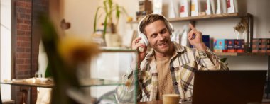 Gülümseyen dijital göçebenin yaşam tarzı portresi, kafede çalışan adam, dizüstü bilgisayarda çalışan, kablosuz kulaklıkla müzik dinleyen, gülümseyen, sandalyede dans eden ve favori şarkının keyfini çıkaran..