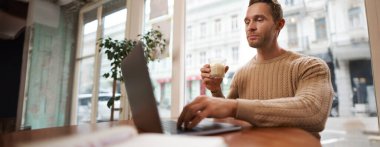 Kafede dizüstü bilgisayarla oturan yakışıklı genç adamın portresi. Dijital göçebe, serbest çalışan, kafede çalışan, bir şeyler yazan..