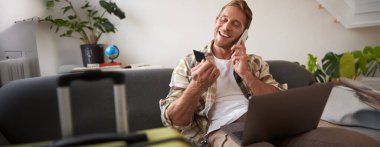 Mutlu turist portresi, dizüstü bilgisayarı ve kredi kartı olan genç bir adam, cep telefonuyla konuşuyor, seyahat acentesini arıyor, tatil için rezervasyon yaptırıyor, yanında bavulu var..