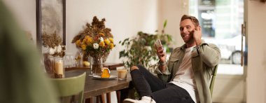 Yakışıklı, şık bir genç adamın portresi, kafeterya ziyaretçisi, akıllı telefonuyla oturuyor ve kablosuz kulaklıkla müzik dinliyor, kameraya gülümsüyor..