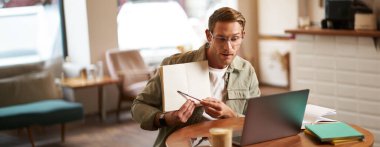 Gözlüklü genç yakışıklı bir adamın portresi, özel ders veren bir öğretmen, defterine işaret eden, bir şeyler gösteren, laptop uygulaması üzerinden video sohbetleri, kafede uzaktan çalışan.