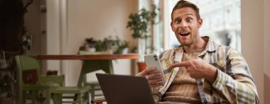 Kafedeki heyecanlı adam, elinde telefonla ekranı göstererek, bir uygulama öneriyor, birlikte çalışma alanında mobil cihaz ve dizüstü bilgisayar kullanıyor. Serbest çalışma alanı ve uzak çalışma alanı kavramı.