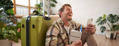 Bavullu yakışıklı bir gencin portresi, yere oturur, akıllı telefona bakar ve güler, pasaport ve tatil için bilet taşır. Turizm konsepti