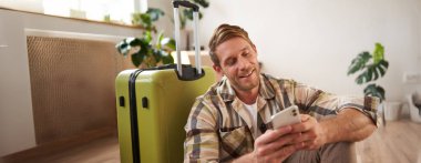 Sosyal medyada gülümseyen yakışıklı bir adamın portresi, akıllı telefondaki mesajla gülüşmeler, mutlu yüzlü cep telefonuna bakmalar, yerde valizin yanında oturmalar..