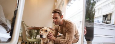 Kafede oturan yakışıklı bir adamın köpeğiyle yakın bir portresi. Bir adam kahvesini içer ve golden retriever 'a dokunur..