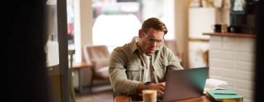 Gözlüklü kızgın bir adamın portresi, hayal kırıklığına uğramış görünüyor ve dizüstü bilgisayarında bir şeyler yazıyor, öfkeli bir yüz ifadesiyle ekrana bakıyor, kafede oturuyor..