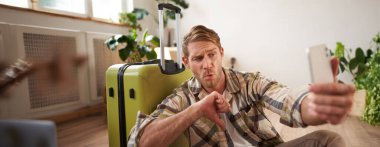 Hayal kırıklığına uğramış bir turist, olumsuz tepkiler veren genç bir adam, baş parmağını kaldırıyor, akıllı telefondan fotoğraf çekiyor, seyahat çantasının yanında oturuyor..