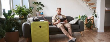 Tabletli genç bir adamın resmi, oturma odasında bavuluyla oturuyor, okuyor, rotayı okuyor, tatile hazırlanıyor. Turizm konsepti