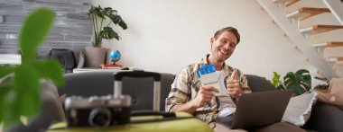 Mutlu yakışıklı adamın portresi, uçuş biletlerini gösteriyor, yaz tatili için yer ayırtıyor, dizüstü bilgisayarla oturma odasında oturuyor, kameraya gülümsüyor..