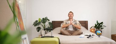 Yolcu yogası portresi, tatile çıkmadan önce meditasyon yapan genç bir adam, bavulla, küreyle ve kamerayla yatak odasında oturuyor, dinleniyor, huzurlu bir tatil arıyor..