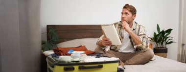 Düşünceli genç bir adamın portresi, tatilini planlayan bir turist, açık bavulla yatakta oturuyor, notlar alıyor, not defterine bir şeyler yazıyor..