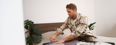 Genç bir adamın portresi, yurtdışına seyahat eden bir turist, bavula giysiler koyuyor, bavulunu kapatıyor, yatağa oturuyor, tatile hazırlanıyor. Turizm konsepti