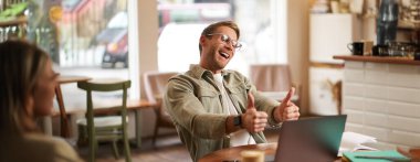 Gözlüklü, mutlu bir gencin portresi, kafede çalışmak, laptopla birlikte çalışmak, onaylamak, iyi olmak, sohbet etmek, online dersler vermek..