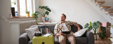 Dijital tabletli genç bir adam, kanepeye oturmuş bavulunu toplamış, ukulele giymiş düşünceli, düşünceli ve tatil planı yapan bir turist. Turizm konsepti