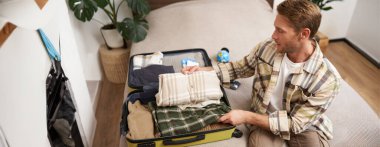 Genç yakışıklı bir adamın portresi, tatile çıkan bir turist, bayram için bavul hazırlamak, bavula giysiler koymak, yatağa oturmak..