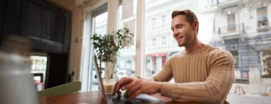 Kafede dizüstü bilgisayarla oturan yakışıklı genç adamın portresi. Dijital göçebe, serbest çalışan, kafede çalışan, bir şeyler yazan..